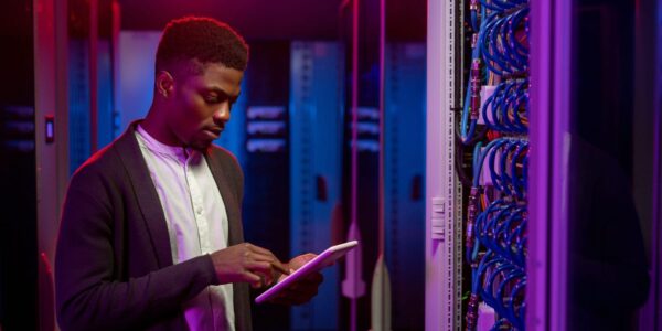 Serious young African-American data engineer standing at open server rack cabinet and using tablet while setting up system at data center